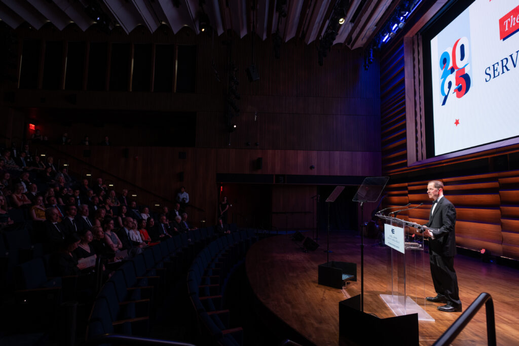 Photo of Max Stier, Partnership for Public Service president and CEO, speaking at the 2025 Samuel J. Heyman Service to America Medals also known as Sammies, from a podium on the right side of the photo to the audience on the left side of the photo